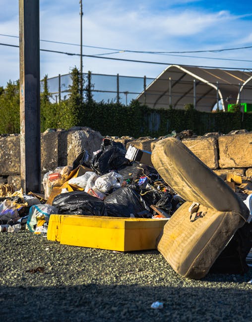 A photograph showing a large pile of mixed household waste on a paved driveway, including cardboard boxes, plastic containers, and discarded furniture, with some items stacked and leaning against each