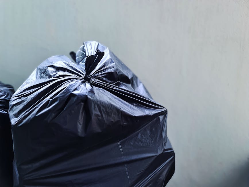 A collection of discarded waste items situated on a rough asphalt surface, including black garbage bags, a yellow plastic storage bin, and an old, dirty car tire leaning against a low stone wall. The 