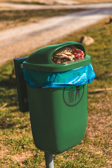 A shoreline scene showing an accumulation of various types of plastic and waste debris, including crumpled black plastic bags, green and brown plastic bottles, clear and colored plastic containers, an